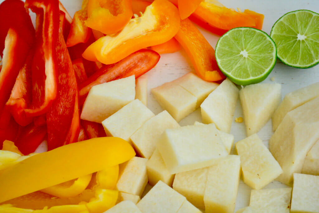 Chopped vegetales sit atop a white cutting board. Red, yellow, and orange bell pepper strips fill the left third of the image while cubed jicama takes up the lower right corner. Peaking out from the top right corner are two halves of a bright green lime.