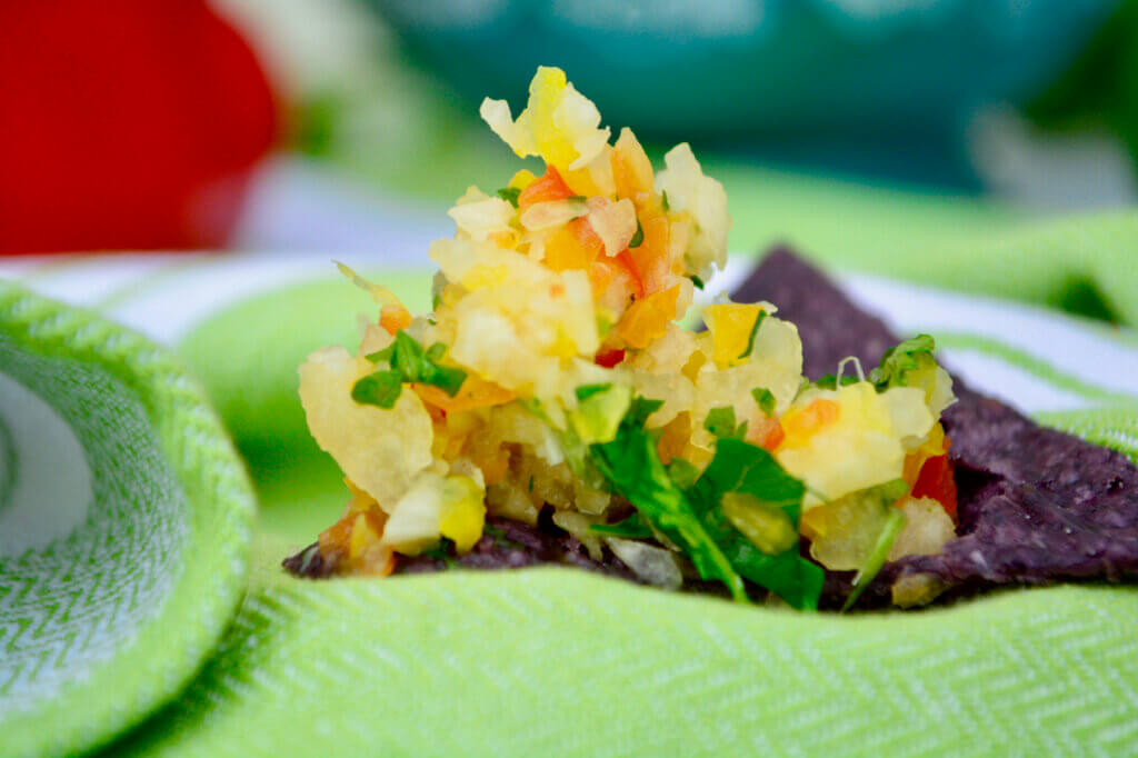 A blue corn tortilla chip sits front and center topped with a heap of multi-colored pico de gallo. A blue patterned bowl and red pepper are blurred in the background with a green and white stripped cloth underlaying all the deliciousness!