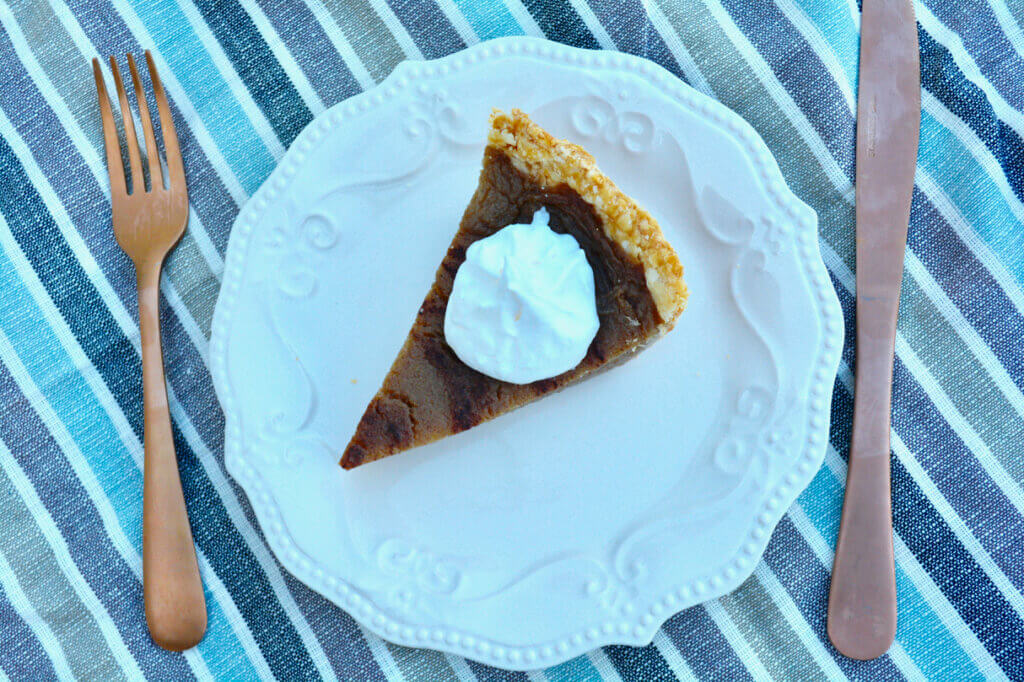 Overhead view of a slice of kidney friendly pumpkin pie made without pumpkin on a white  plate with a fork and knife next to it on a blue striped tablecloth.