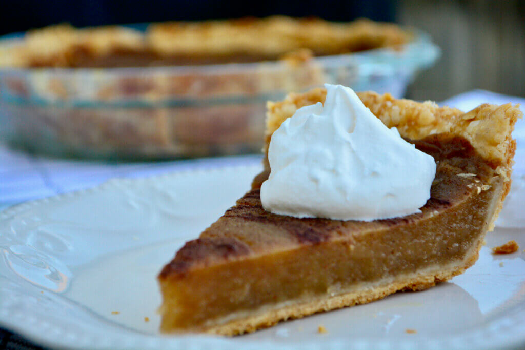Close up of a slice of kidney friendly pumpkin pie on a white plate with a dollop of whipped cream on top with entire pie in the background on a glass plate