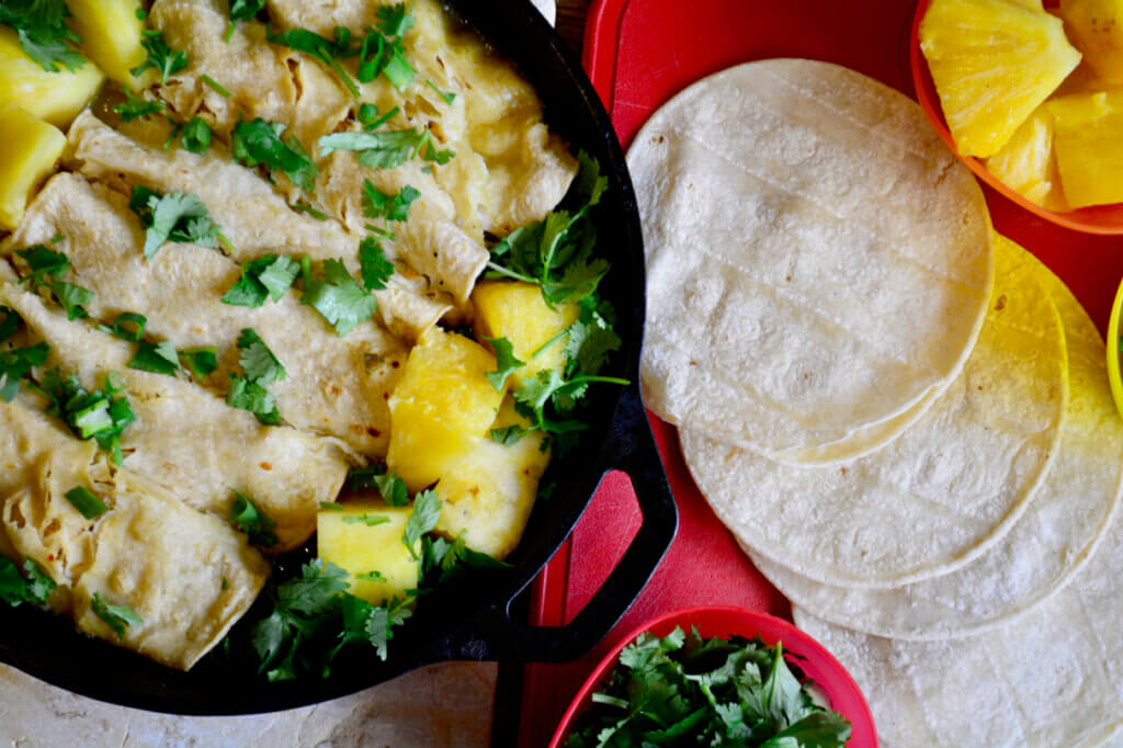 A warm skillet of pineapple enchiladas is featured in the top left corner surrounded by cilantro and tortillas.