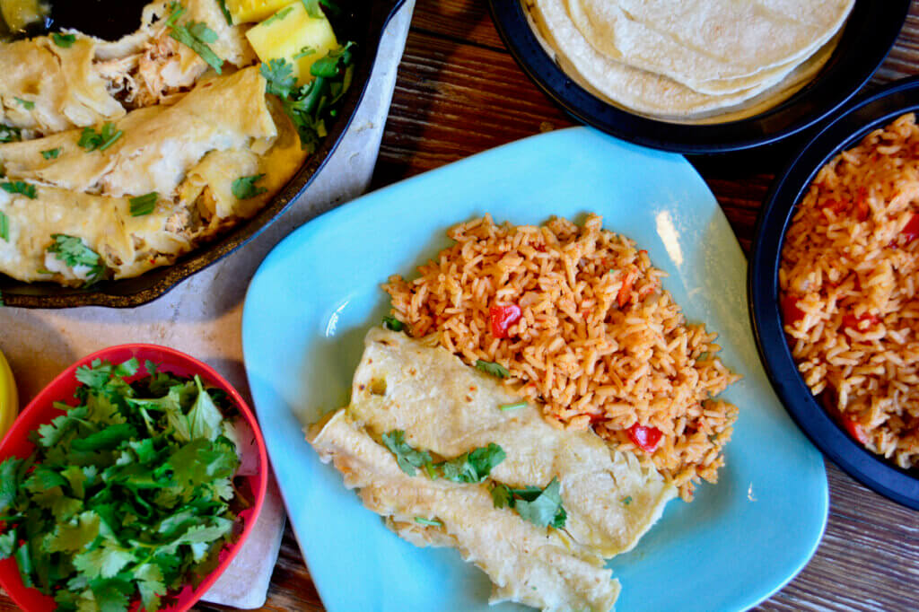A turquoise plate of two pineapple enchiladas and a side of Spanish rice is featured in the bottom left. Tortilla's, cilantro, and a skillet full of enchiladas surrounds the brightly colored plate.