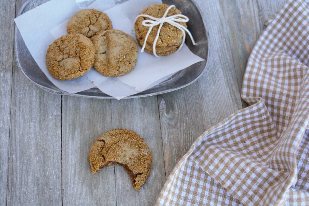 Overhead view of kidney friendly ginger cookies on parchment in a silver oval bowl with a gingham tea towel next to it and one cookie with a bit taken out of it