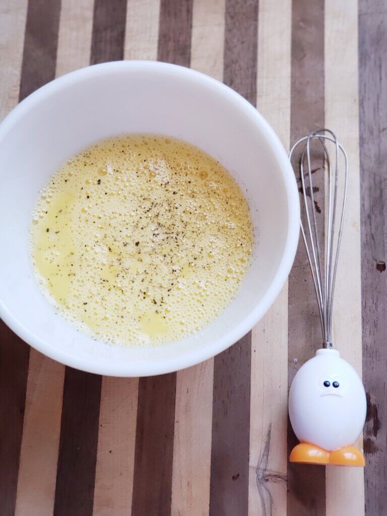 Beaten eggs in a white bowl with an egg whisk sitting next to it on a striped cloth