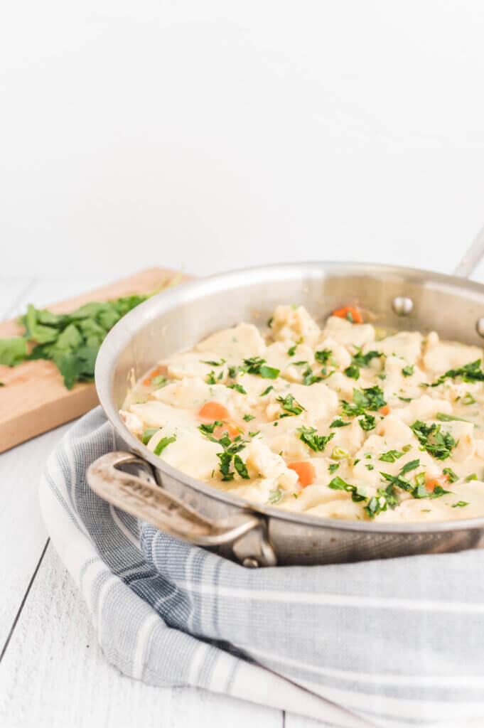 Chicken and dumplings in a stainless steel sauté pan with a wooden cutting board with fresh parsley in the background