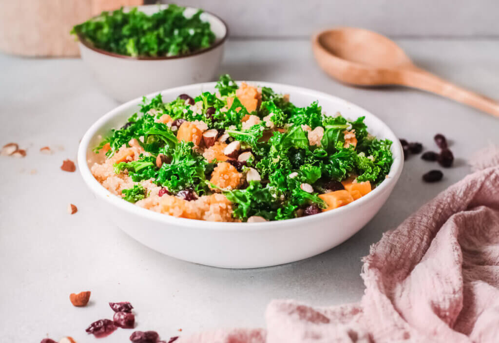 Cranberry quinoa sweet potato salad side view with bowl of greens and wooden spoon. plant-based