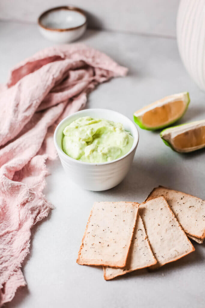 Avocado crema in a white bowl side angle with pink dish towel, lemon and crackers