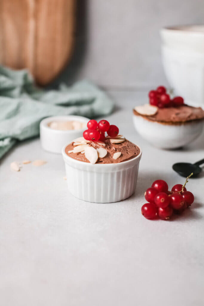 Side view of Coconut chocolate almond mousse in a white ramekin with berries and small white bowl of mousse in the background with green towel and wooden board