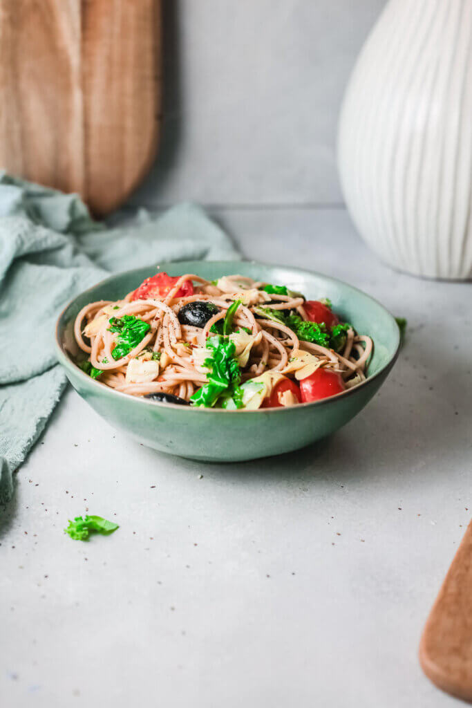 Side angle view Mediterranean pasta with kale in a teal bowl with blue dish towel, wooden board and white vase in background
