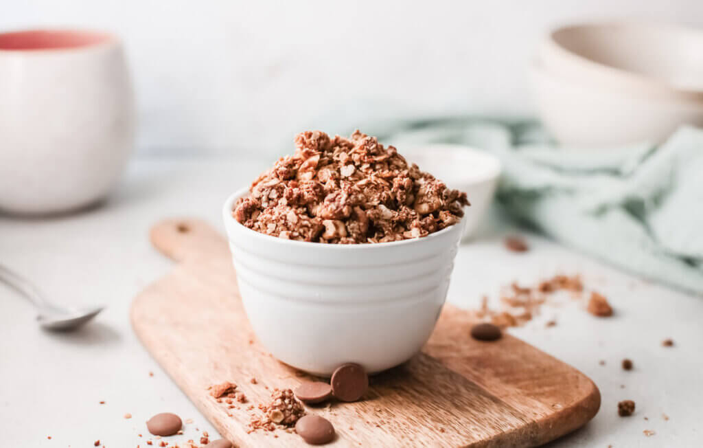Side view of lightly salted dark chocolate pecan granola in a small white bowl on a wooden board with green towel and small white bowls