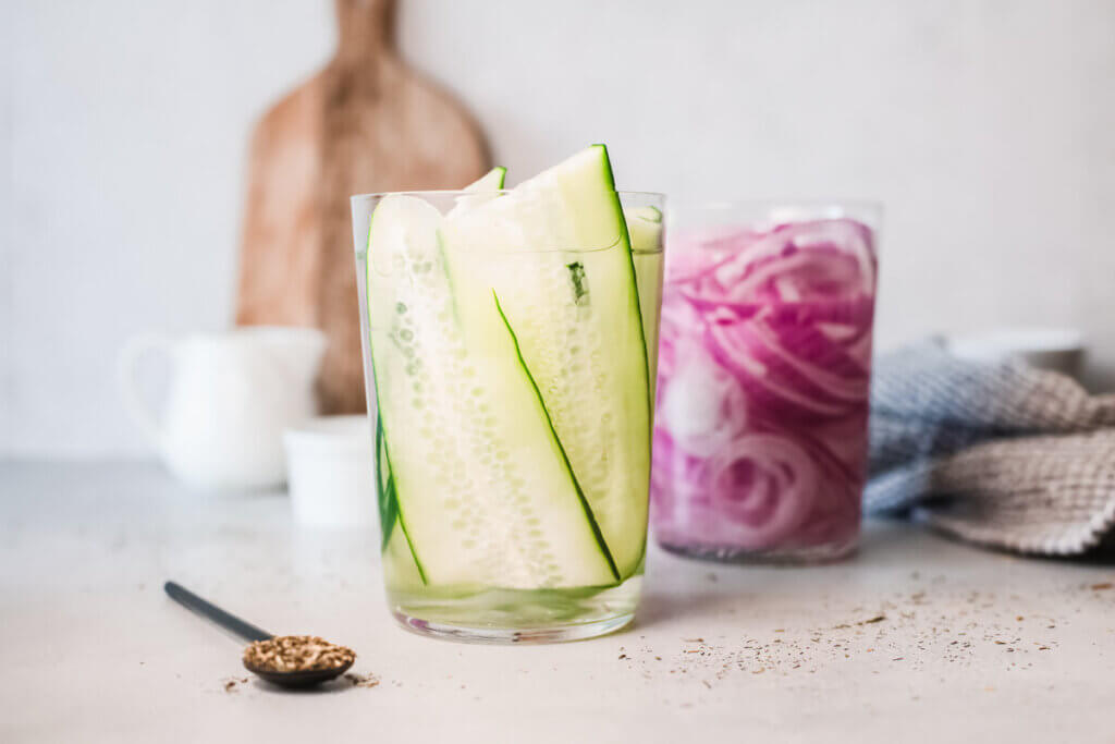 Low sodium quick pickled cucumbers and onions in glass with white bowls and a spoon and wooden board in the background