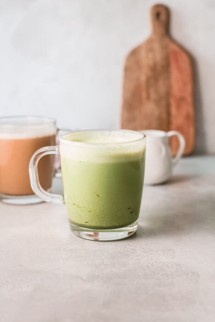 Side view of Coconut milk matcha latte in a clear mug with a clear mug of coconut milk latte and white small pitcher and wooden board in the background