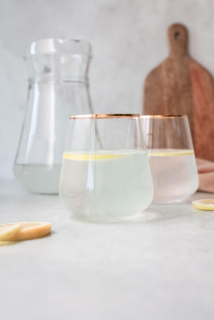 Side view keto kidney electrolyte replacement drink in two glasses with gold trim, large water carafe, lemon slices, pink tea towel and wooden cutting board standing in background