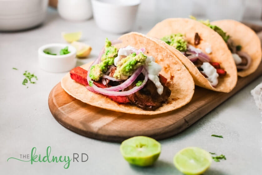 Side view of three marinated mushroom and carrot tacos on a wooden board with lime sliced in half and white bowls in the background
