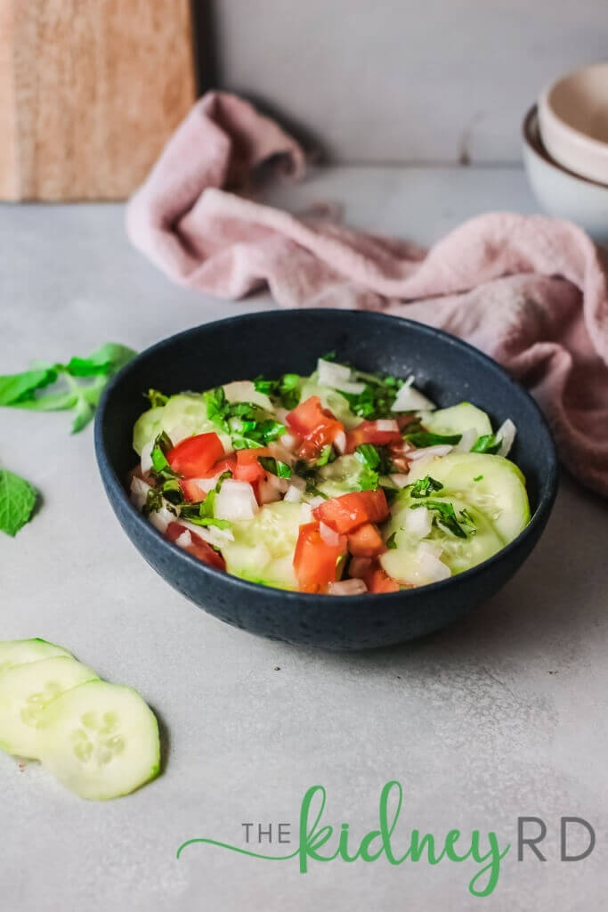Zoomed out view of Mediterranean cucumber salad in a black bowl with cucumber slices and pink tea towel with wooden board in background and white bowls