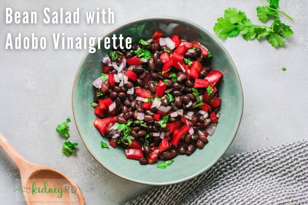 Overhead view of make ahead bean salad with adobo vinaigrette in a blue bowl with a wooden spoon, grey tea towel and cilantro sprig in the background.