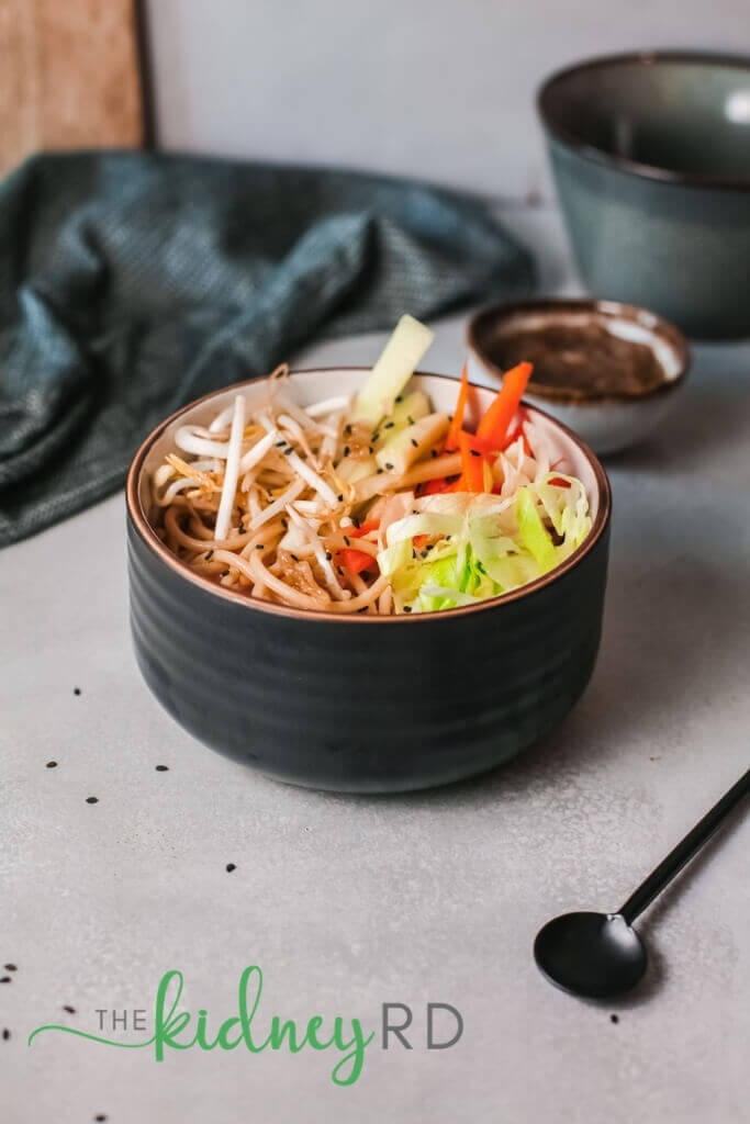 Side view of cold vegan asian noodle salad in a black bowl with a black spoon and green dish towel in background