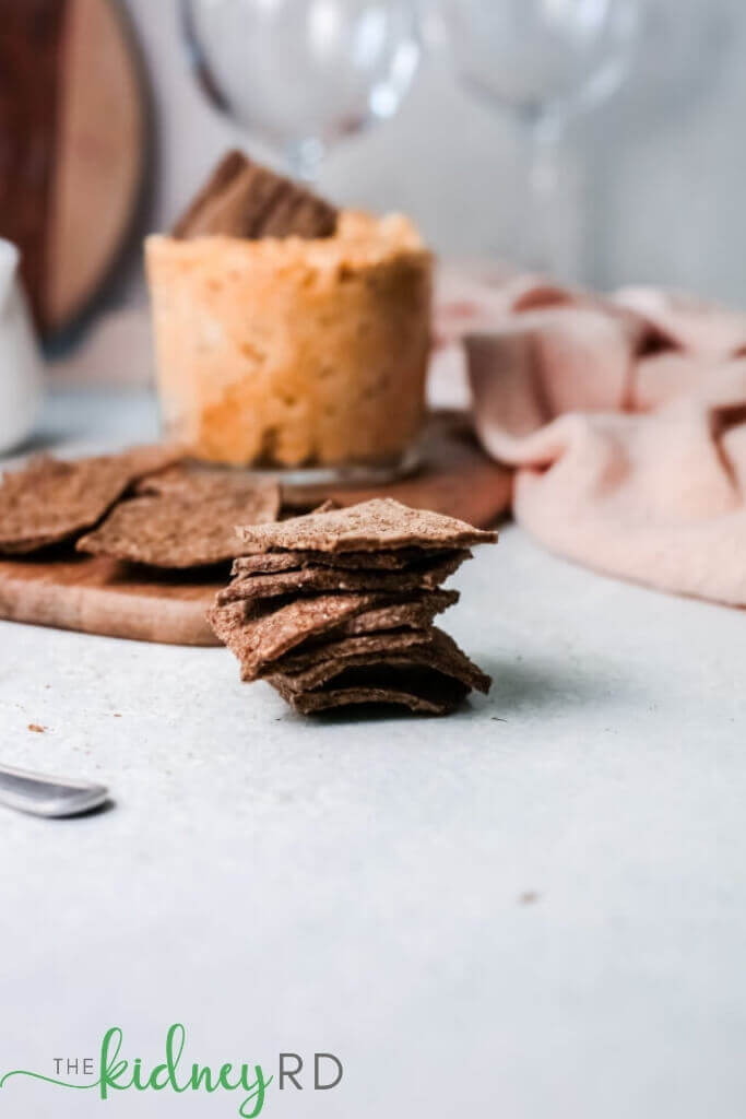 Stack of keto flax crackers with a wooden board with more flax crackers and artichoke hummus in a glass bowl, pink tea towel and 2 wine glasses in the background