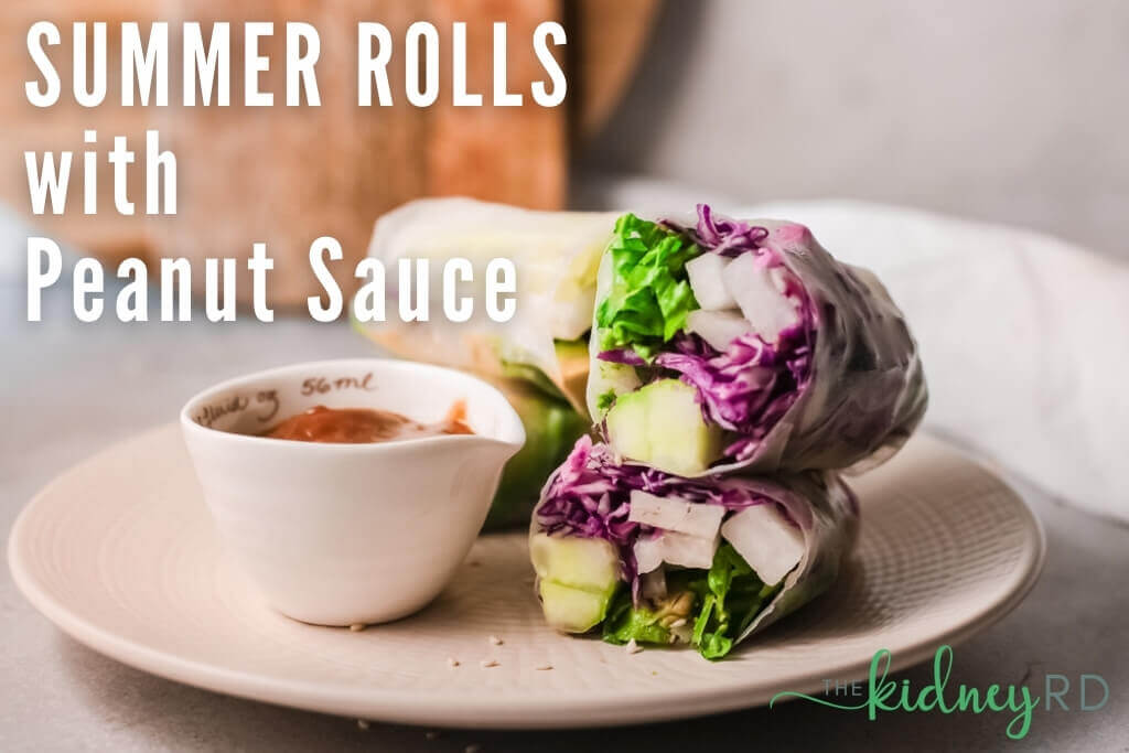 Side view of summer rolls and small white bowl with peanut sauce on a white plate with wooden cutting boards in the background