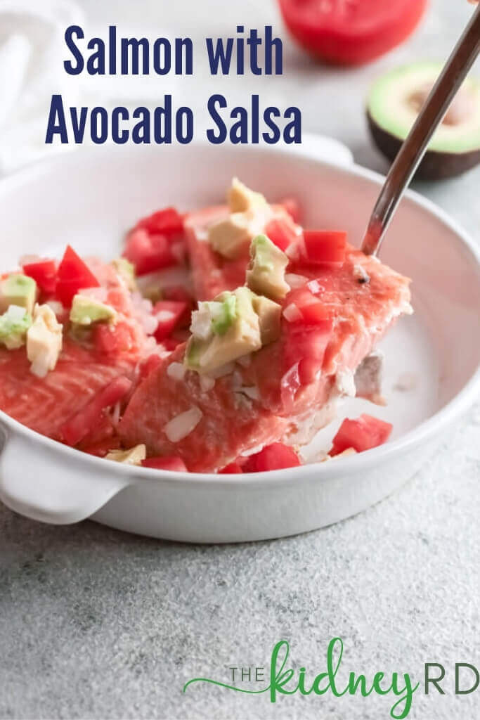 Close up view of keto salmon with avocado salsa on a white plate with a fork breaking off a piece. Half of an avocado and tomato in the background