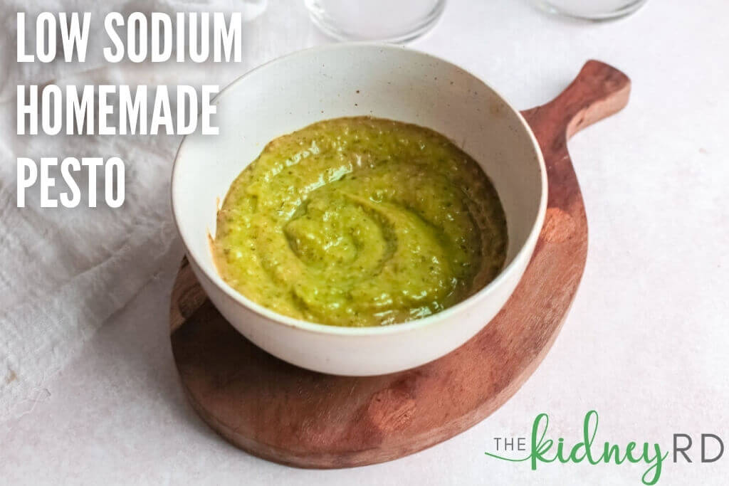 Low sodium homemade pesto in a white bowl on a wooden board and white tea towel and water glasses in the background