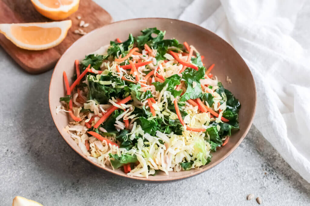 Kale citrus slaw in a beige bowl with white tea towel and lemon wedges on a cutting board next to it