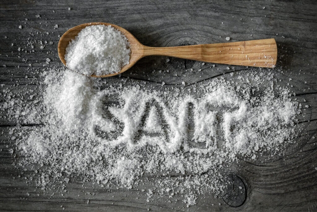 Overhead view of salt on a wooden spoon with the word salt written in spilled salt next to spoon