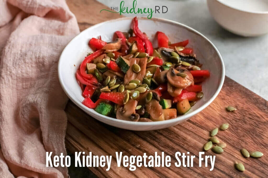 Keto kidney vegetable stir fry in a white bowl sitting on a wooden board with a pink tea towel, pumpkin seeds next to bowl and white bowl in background
