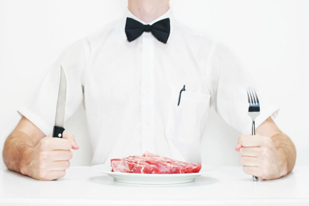 Man in a white short-sleeved button up and black tuxedo  tie with a fork and knife and large plate of raw steak on a white plate