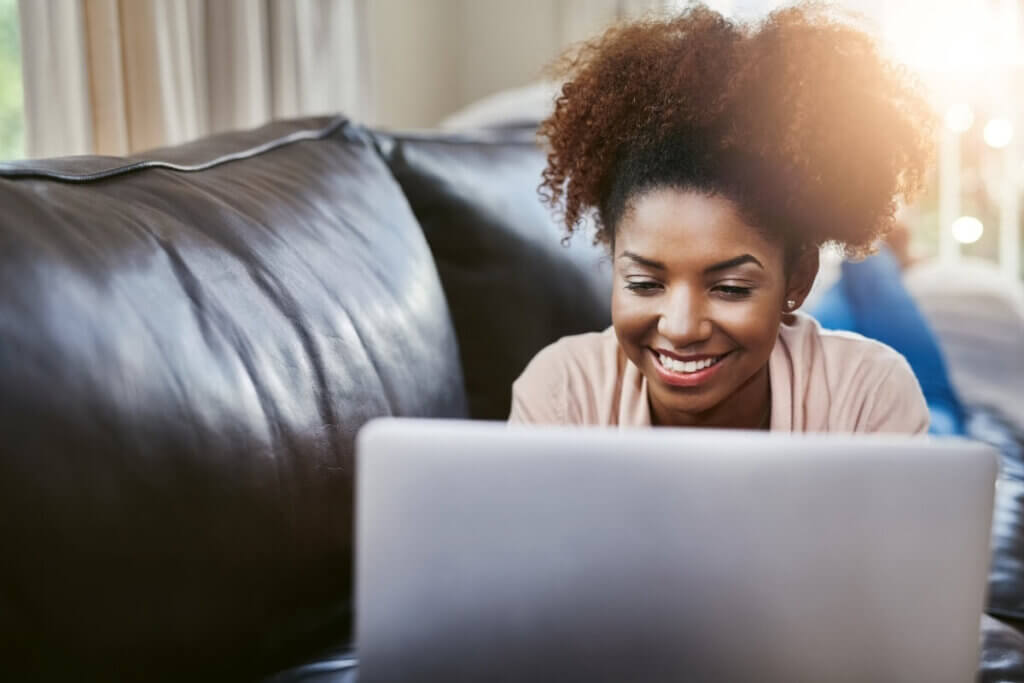 Young woman of color on a brown leather couch smiling at her open laptop