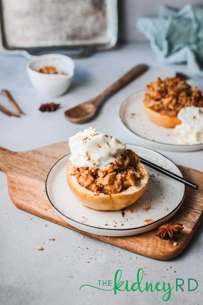 Low potassium dessert of baked apple with whipped cream on a white plate sitting on a wooden board with another dish of baked apple a wooden spoon, green dish towel and sheet pan in the background