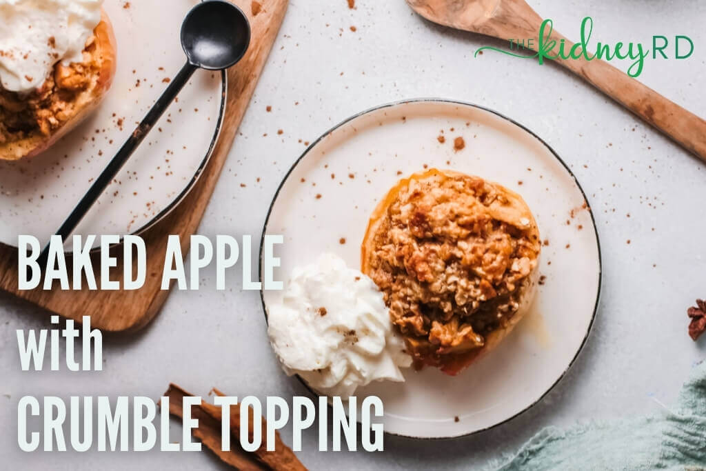Top view of low potassium dessert of baked apple with crumb topping on white plate with dollop of cream with wooden spoon cinnamon sticks and green dish towel next to it