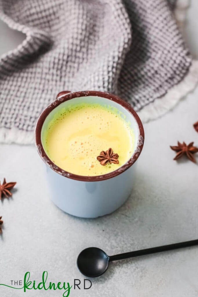 Side view of kidney friendly golden milk, a hot drink for kidney health, in a white mug with a black spoon and grey tea towel with flower shaped small ginger cookies
