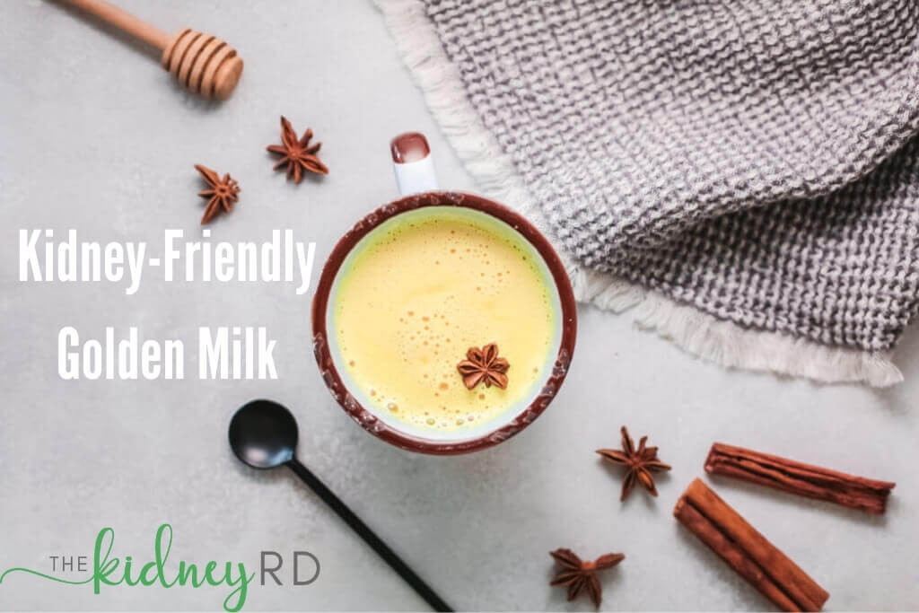 Overhead view of kidney friendly golden milk in a white mug with brown border surrounded by a honey spoon, cinnamon sticks, a small black spoon and a grey tea towel.