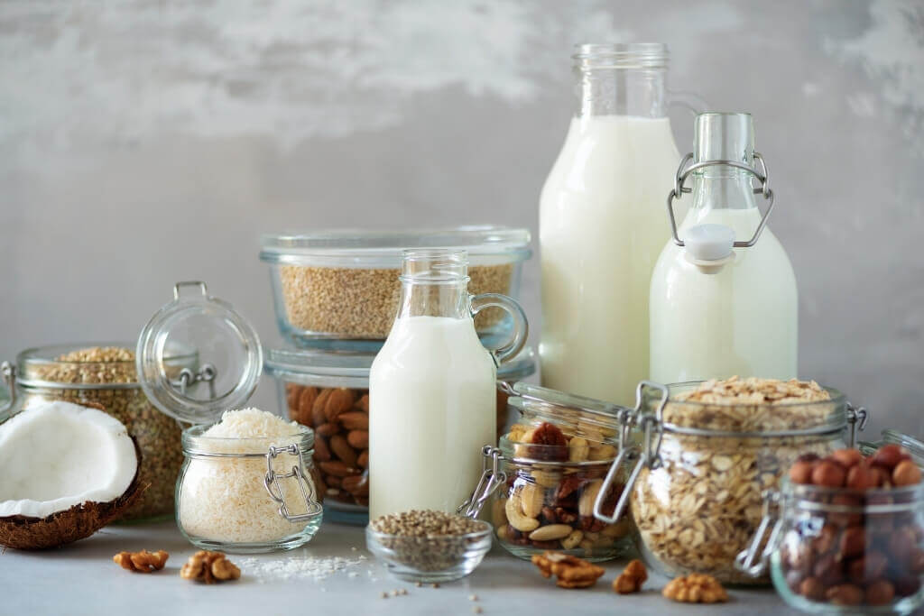 Glass mason jars filled with different nuts and seeds and three large glass bottles of plant-based milk
