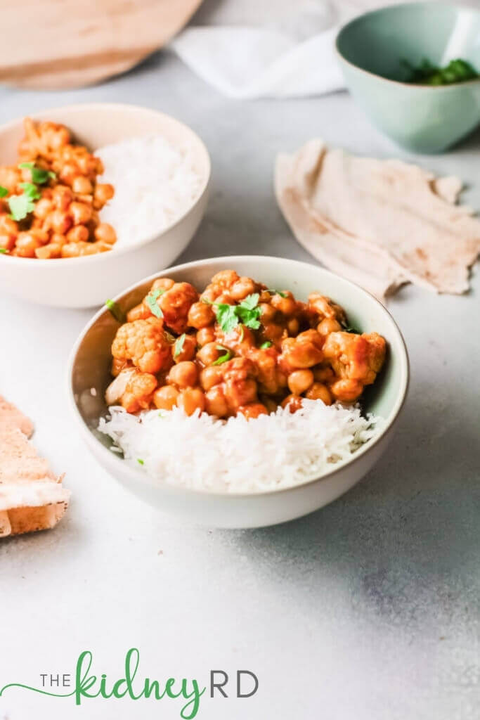 Close up view of vegan tikka masala in a green bowl with white rice and pita bread halves with a chopped cilantro in a green bowl in the background