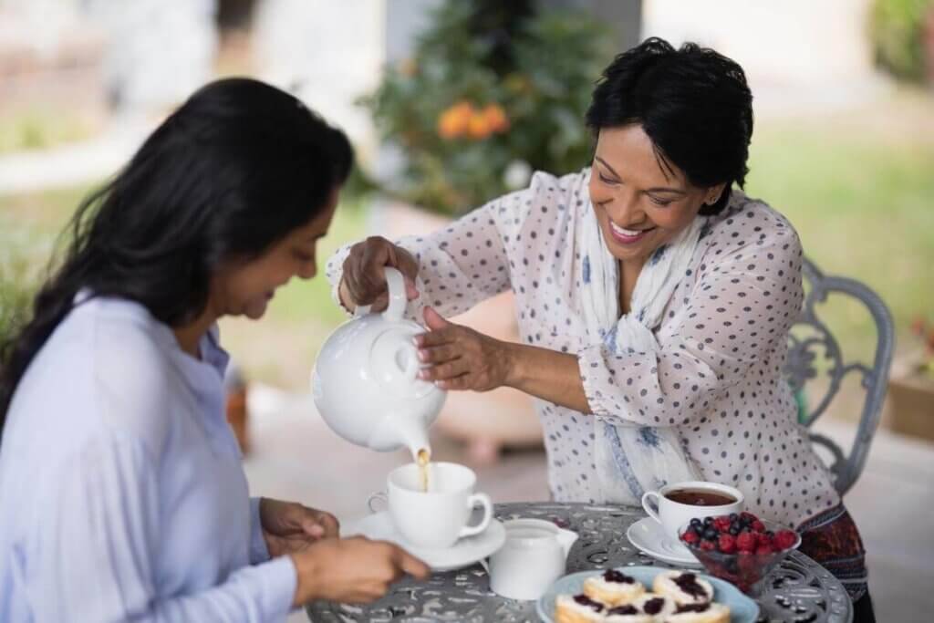 Two smiling women sitting at a small table pouring tea from a white ceramic tea pot into a white tea cup with saucer and a bowl of fruit on the table.