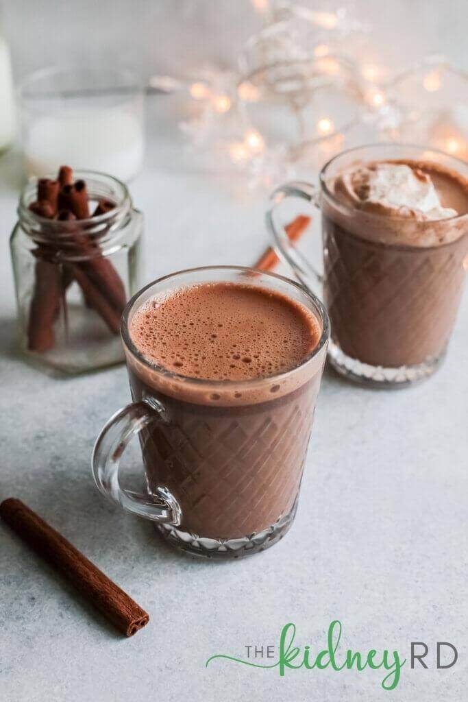 Overhead angled view of 2 glass mugs of spiced hot mocha keto with cinnamon sticks and a string of twinkle lights in the background