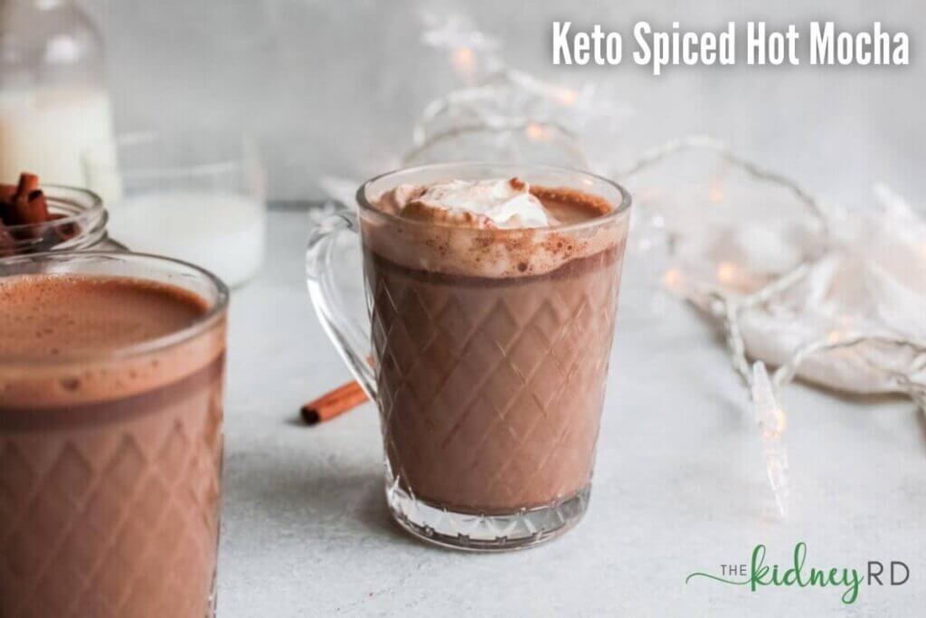 Side view of keto spiced mocha in a glass mug with whipped topping and a string of lights, white tea towel and cinnamon sticks in the background