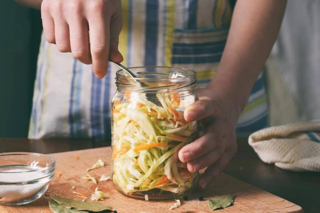 Woman  with a striped apron making sauerkraut in a mason jar sitting on wooden cutting board with a bowl of sugar and bay leaves on cutting board
