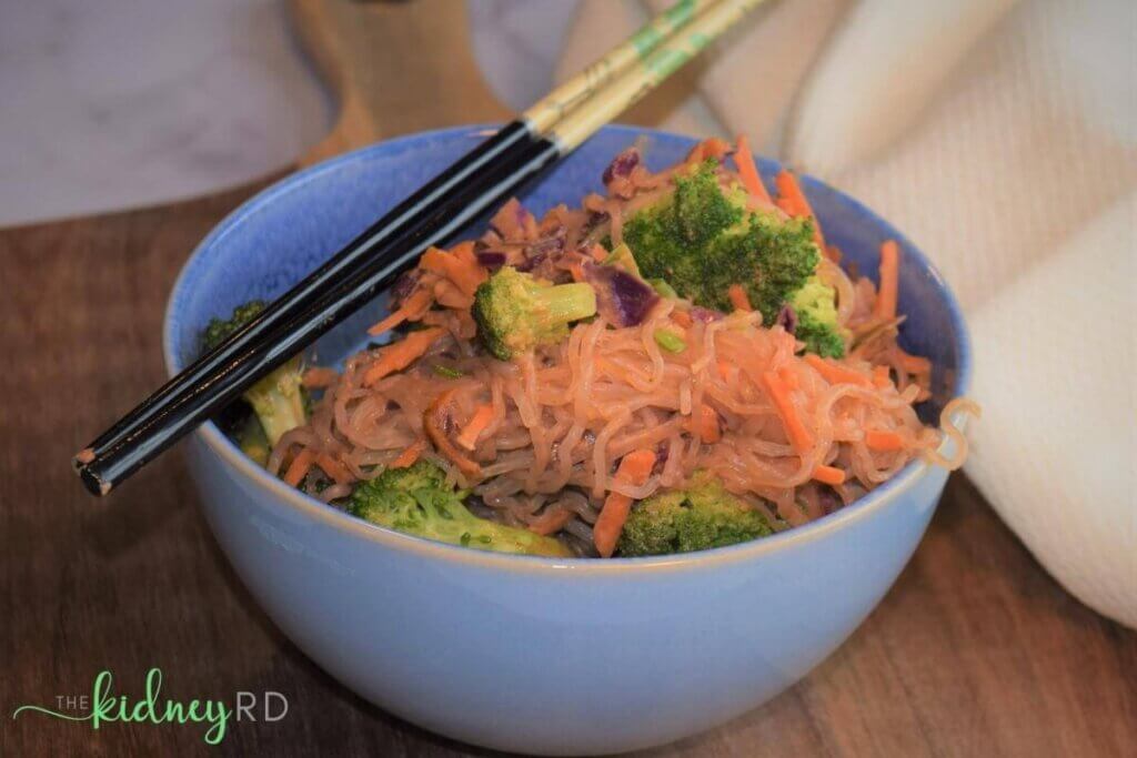 Side view of shirataki noodle stir fry in a blue bowl with black chop sticks laying across bowl on a wooden board with a cream  towel next to it