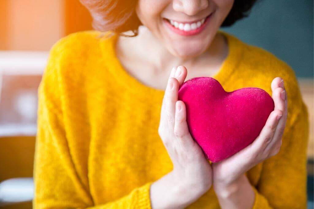 Smiling woman in a yellow sweater holding a red heart in her hands