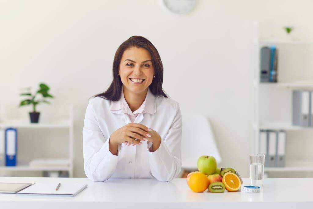 Smiling dietitian sitting at a desk with a pad of paper and fruits and a glass of water next to her
