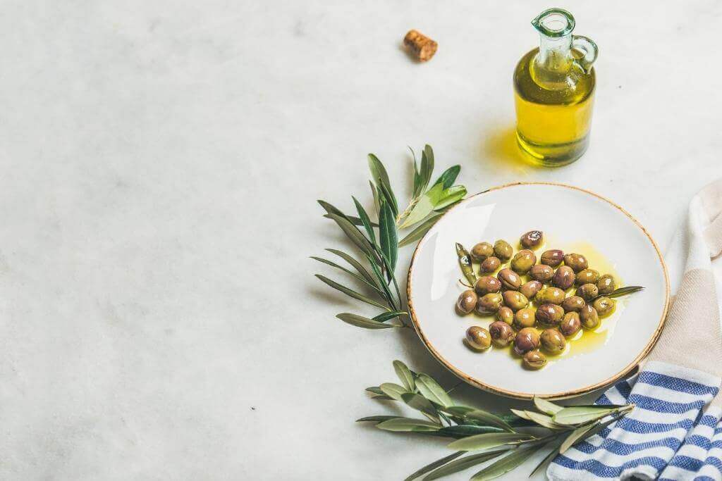 Picture of olives on a white plate drizzled in oil with olive leaves and a blue and white tea towel next to it along with a glass bottle with olive oil