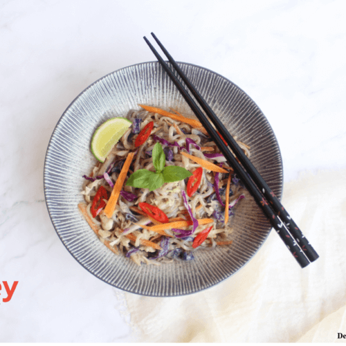 A bowl of Thai curry shirataki noodle stir fry with shredded green cabbage, purple cabbage, and carrot with a garnish of fresh basil and a wedge of lime
