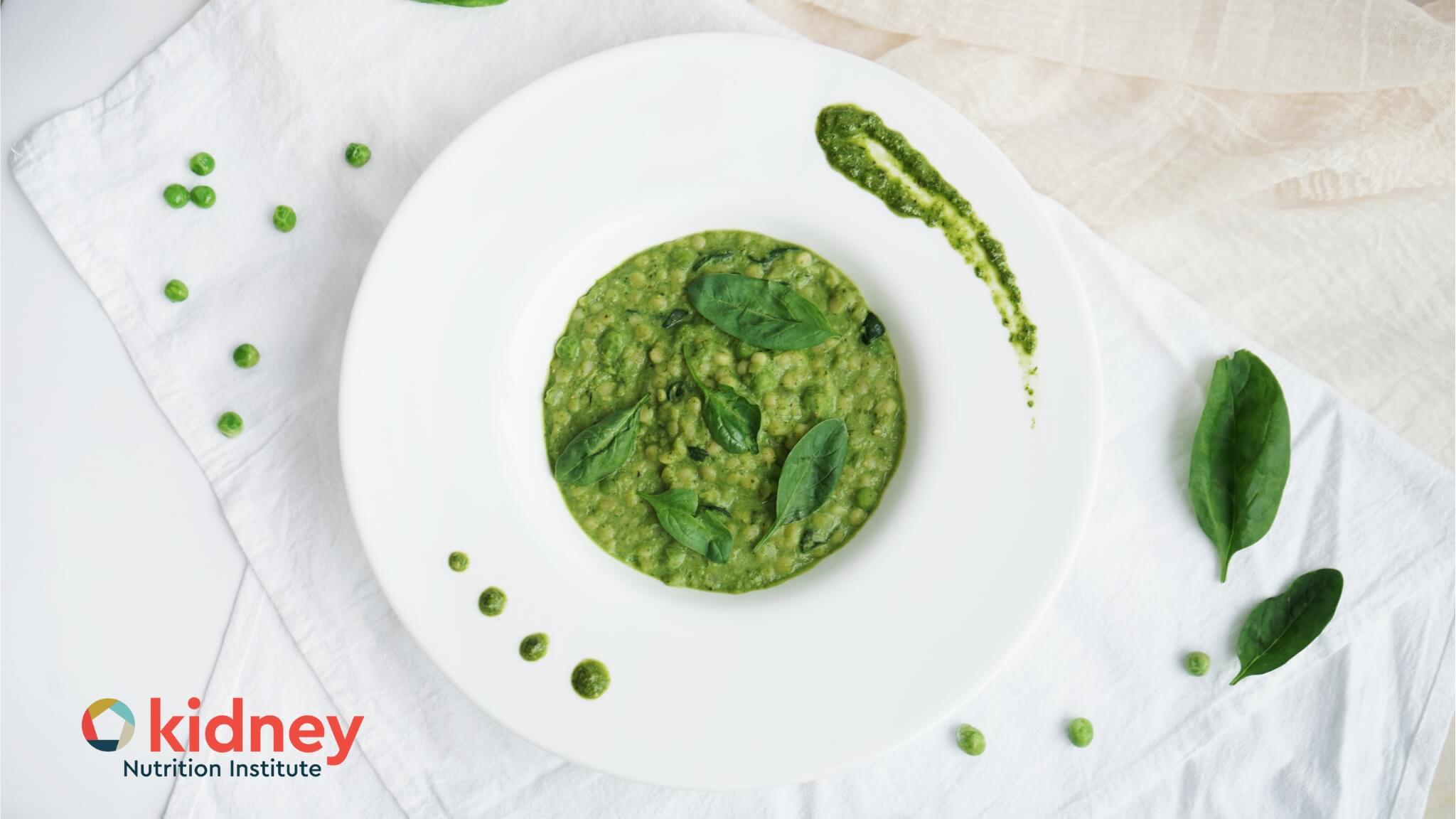 Green creamy spinach couscous risotto in a white pasta plate with a streak of pesto, spinach leaves, and peas around the edges