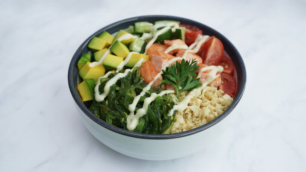 A salmon poke bowl with cauliflower rice, salmon, assorted vegetables, and spicy mayonnaise drizzled as garnish