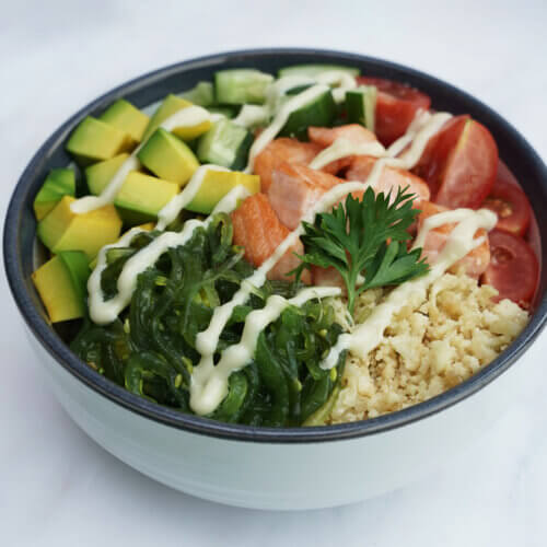 A salmon poke bowl with cauliflower rice, salmon, assorted vegetables, and spicy mayonnaise drizzled as garnish